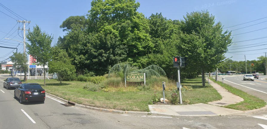 Daytime street view showing cars driving past a landscaped corner with a traffic signal and a wooden "Welcome to Coram" community sign.