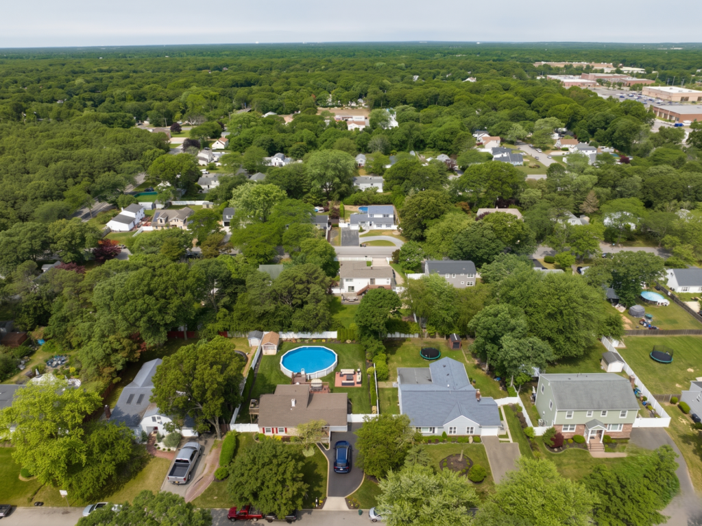 Aerial view of a lush suburban neighborhood with backyard pools and tree-lined streets stretching toward a dense forested horizon in Medford, New York.
