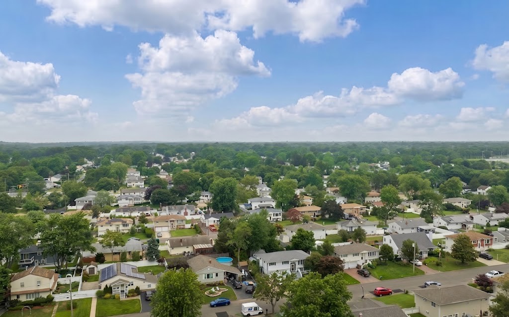 Aerial view of a sprawling, tree-filled suburban neighborhood featuring residential homes, a distant water tower on the horizon, and a bright, cloud-dotted sky in Selden, New York.