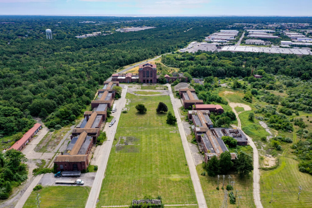 Aerial view of the historic Pilgrim State Psychiatric Center campus in Brentwood, New York, surrounded by dense green forest, commercial buildings, and a water tower in the distance.