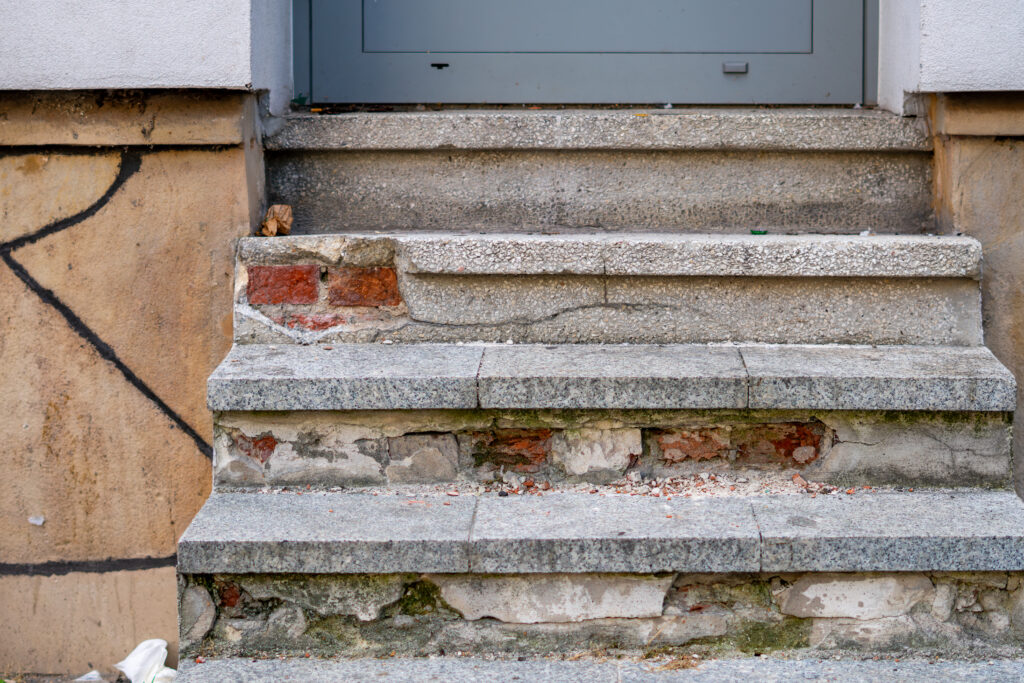 A close-up view of severely cracked brick and stone stairs at a residential house entrance, illustrating a hazardous design defect and property maintenance negligence.